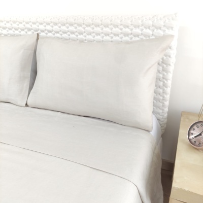 Cama com almofadas e lençol bege claro, cabeceira branca e mesa de cabeceira com relógio.