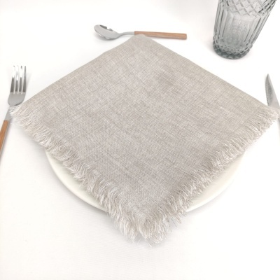 Guardanapo cinza claro com franjas sobre prato branco, utensílios de mesa com cabo de madeira e copo de vidro texturizado