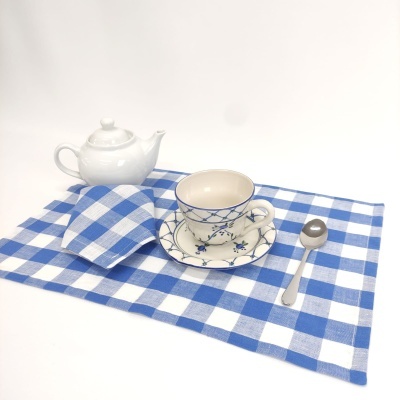 Conjunto de chá com bule branco, chávena e prato com padrão azul, guardanapo e toalha xadrez azul e branco, colher de metal