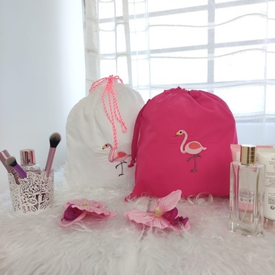 Dois sacos de tecido com bordado de flamingo, pincéis de maquilhagem, flores artificiais e frascos de perfume sobre superfície branca peluda