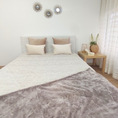 Cama com cabeceira trançada branca, colcha branca e manta cinza, almofadas bege, mesa de cabeceira com vaso, livro e candeeiro