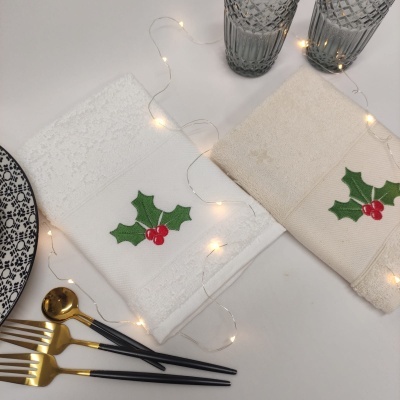 Dois guardanapos bordados com folhas de azevinho e bagas vermelhas, com talheres dourados, copos de vidro e prato decorado ao lado