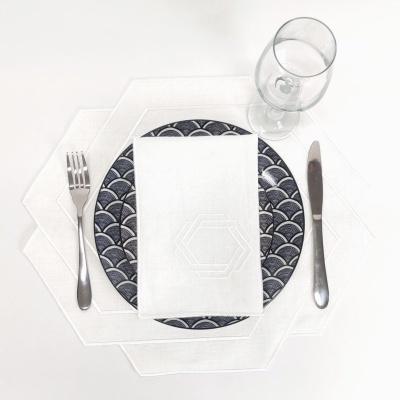 Conjunto de jantar com prato azul e branco, guardanapo branco, talheres e taça sobre jogo americano branco hexagonal.
