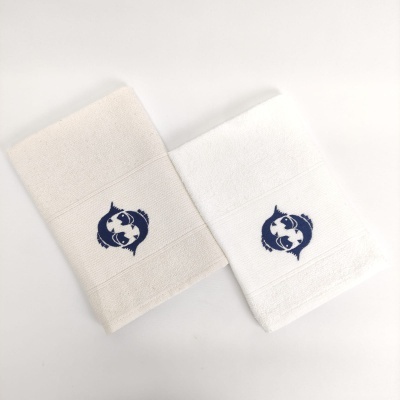 Duas toalhas texturizadas, uma branca e uma bege, com bordado de peixes azuis entrelaçados.