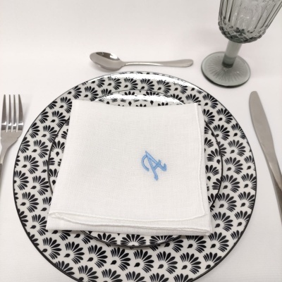 Prato decorativo branco e preto com guardanapo branco monogramado azul, talheres metálicos e copo de vidro cinza texturizado.