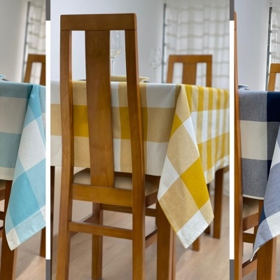 Três toalhas de mesa xadrez em azul claro, amarelo e azul escuro em mesas de madeira com cadeiras, cortinas brancas ao fundo.