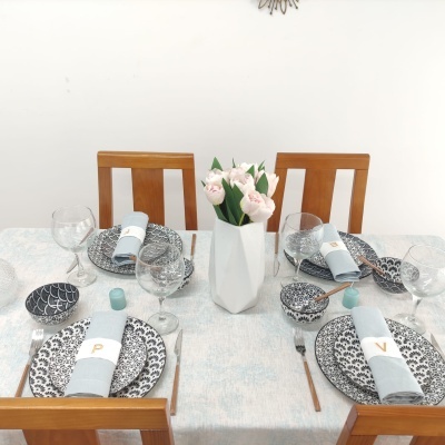 Mesa de jantar posta com pratos cerâmicos preto e branco, guardanapos cinzentos, talheres de madeira e vaso branco com flores cor-de-rosa