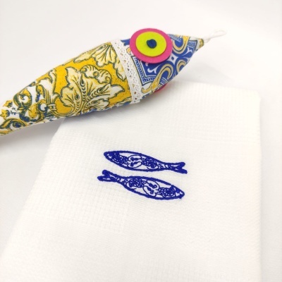 Saco decorativo em tecido amarelo com padrão colorido sobre toalha branca bordada com peixes azuis.