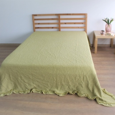 Cama com colcha verde claro e cabeceira de madeira clara junto a mesa de cabeceira com vaso de flores