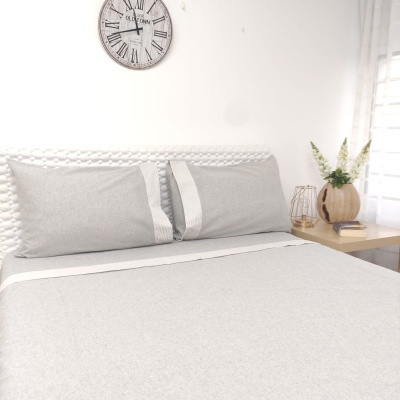 Roupa de cama cinza claro com detalhes brancos em cama com cabeceira branca, mesa com vaso, livros, castiçal e relógio de parede