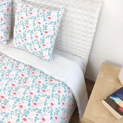 Conjunto de cama com padrão floral em rosa e verde, cabeceira branca e mesa de cabeceira com livros