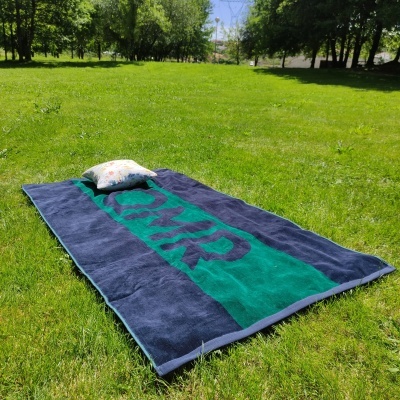Toalha de praia azul escuro e verde com letras AUP numa relva verde com almofada
