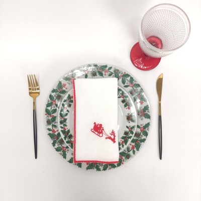 Mesa posta com prato natalício, guardanapo bordado, talheres dourados e copo plástico vermelho e transparente