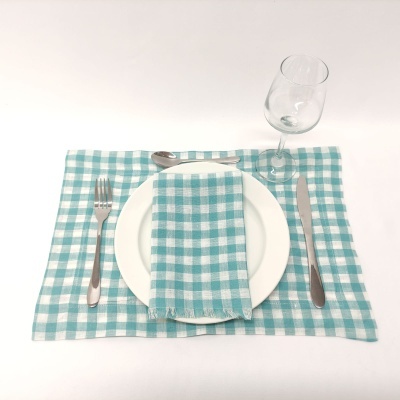 Mesa posta com jogo americano e guardanapo xadrez azul e branco, prato branco, talheres inox e copo de vidro