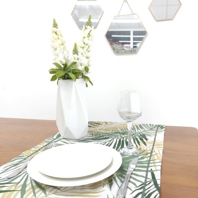 Mesa com toalha de padrão folhagem, jarro branco com flores, pratos brancos, faca e copo de vidro