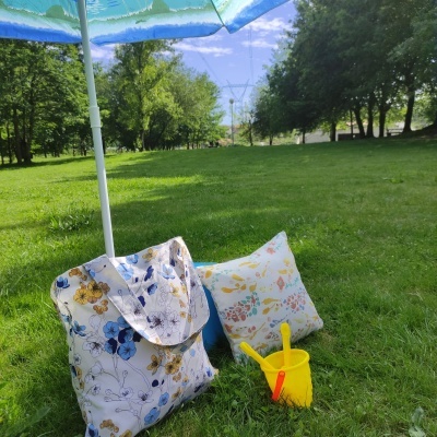 Guarda-sol, mala floral, almofada floral, balde amarelo e pá vermelha num relvado verde e árvores ao fundo