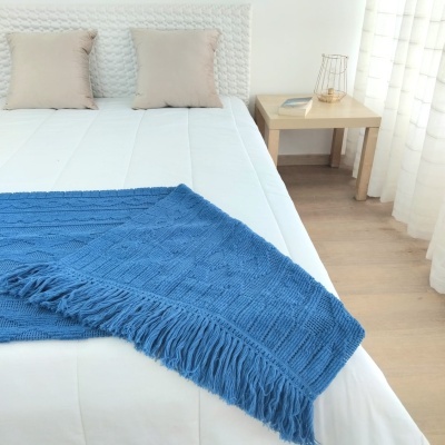 Cobertor azul de malha com franjas numa cama branca com almofadas bege e mesa de cabeceira