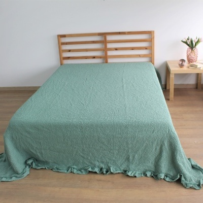Cama com colcha verde água texturizada e cabeceira de madeira, mesa lateral com vaso de flores.