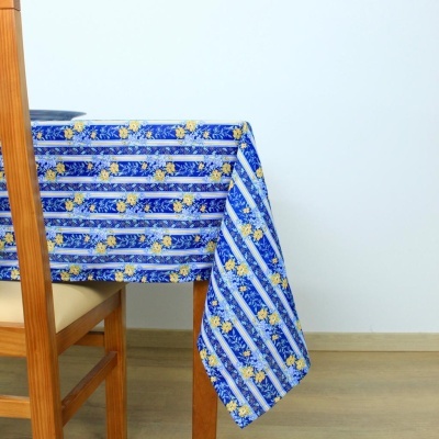 Toalha de mesa azul com flores amarelas e riscas brancas numa mesa de madeira clara com cadeira ao lado.