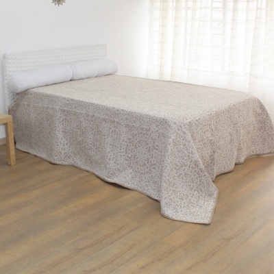 Cama com colcha bege floral, cabeceira branca e almofadas cilíndricas brancas perto de janela com cortinas brancas