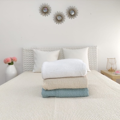 Três mantas dobradas em branco, bege e azul claro sobre cama branca texturizada com almofadas e decoração de espelhos na parede