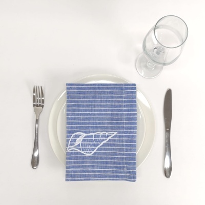 Mesa posta com guardanapo azul às riscas e desenho de croissant, prato branco, talheres de metal e copo de vidro