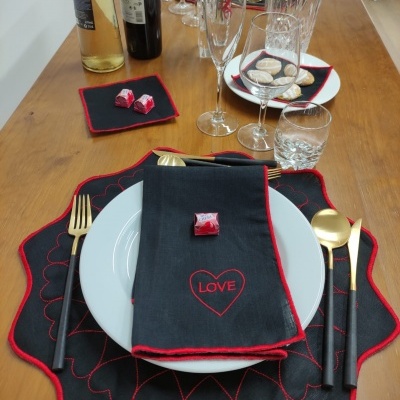 Mesa posta com prato branco, individual preto com borda vermelha e padrão de coração, guardanapo preto com coração LOVE, talheres dourados e copos de vidro