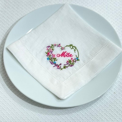 Guardanapo branco com bordado colorido e palavra 'Mãe' dobrado sobre prato branco