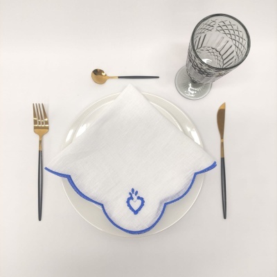 Mesa posta com prato branco, guardanapo branco com bordado azul, talheres pretos e dourados, colher e copo cinzento