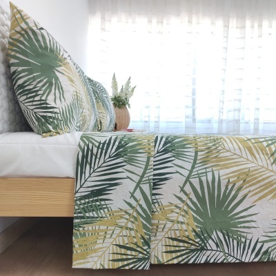 Cama com roupa de cama estampada com folhas verde e bege ao lado de janela com cortinas brancas e planta decorativa