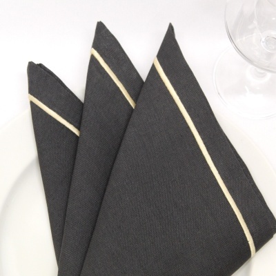 Guardanapo cinzento escuro com risca bege dobrado sobre prato branco e copo de vidro