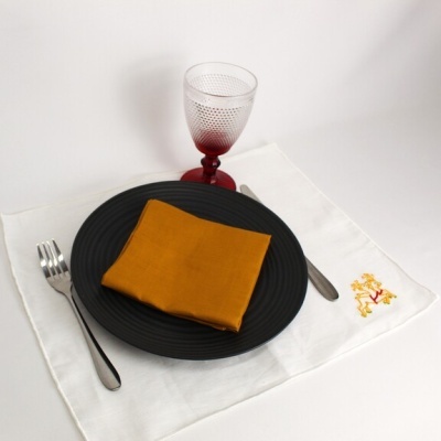 Mesa posta com prato preto, guardanapo mostarda, talheres de aço inox, copo vermelho e toalhete branco bordado