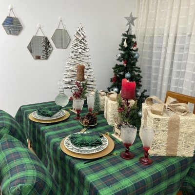 Mesa de jantar decorada com toalha xadrez verde e azul, pratos e copos com velas e árvores de Natal