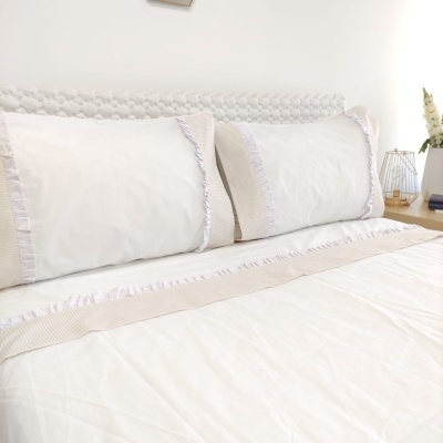 Cama com fronhas e lençol branco com detalhes bege e cabeceira cinza claro em tecido trançado