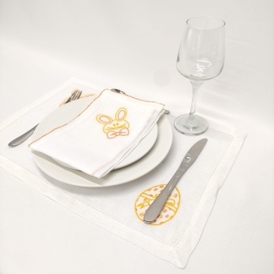 placemat branco com bordado amarelo, guardanapo branco com bordado de coelho amarelo, prato branco, talheres metálicos e copo de vidro transparente