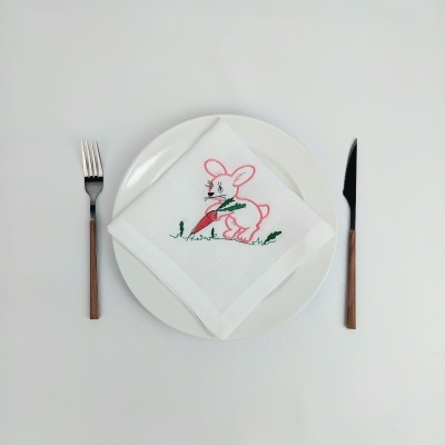 Guardanapo branco bordado com coelho rosa sobre prato branco, garfo e faca com cabo de madeira ao lado
