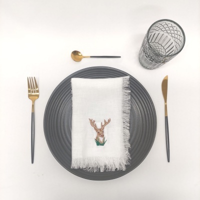 Mesa posta com prato cinzento, guardanapo branco bordado com veado, talheres preto e dourado, colher pequena dourada e copo transparente