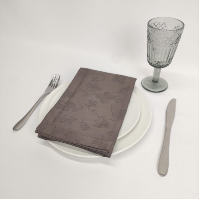 Conjunto de mesa com prato branco, guardanapo castanho com padrão floral, talheres prateados e copo de vidro texturizado