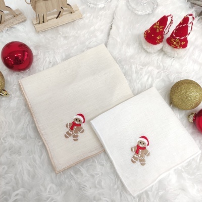Dois panos de cozinha com bordados de biscoitos de gengibre com gorros de Natal sobre uma superfície peluda branca, com bolas e botas decorativas de Natal ao redor.
