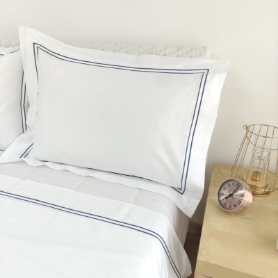 Roupa de cama branca com linhas azuis, despertador cobre e candeeiro dourado numa mesa de madeira