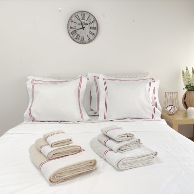 Conjunto de roupa de cama branca com linhas vermelhas, toalhas bege e cinza em cama branca, relógio e decoração na parede branca