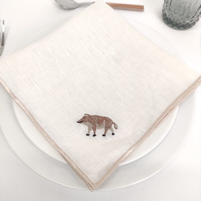 Guardanapo branco de tecido com bordado de raposa castanha sobre prato branco numa mesa com talheres e copo cinzento