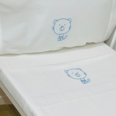 Jogo de cama branco com bordados azuis de animalzinho, almofada e lençol numa cama branca.