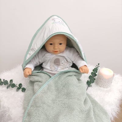 Boneco bebé com toalha com capuz verde esverdeado e body cinza sentado sobre tecido branco felpudo