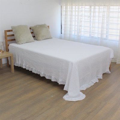 Cama com colcha branca texturizada e almofadas bege, em quarto com piso e móvel de madeira clara