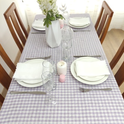 Mesa de jantar com toalha xadrez lilás e branco, pratos, talheres, guardanapos, velas e vaso com flores brancas