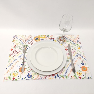 Conjunto de prato branco, talheres e taça sobre individual com design floral e texto colorido.