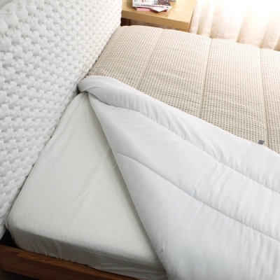 Cama com colchão branco, protector branco, manta branca, colchão beje com padrão, cabeceira branca texturizada e mesa de cabeceira com livros junto à janela