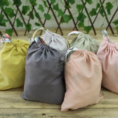Seis sacos de tecido coloridos com cordão, sobre mesa de madeira e fundo de plantas