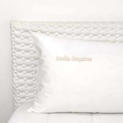 Travesseiro branco bordado com texto 'Amélia Gonçalves' e cabeceira branca texturizada